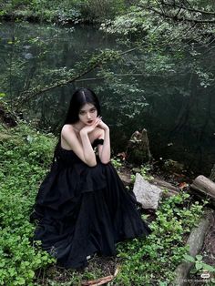 a woman in a black dress sitting on the ground next to a body of water