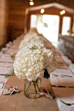 white flowers are in a vase on a long table with other place settings and napkins