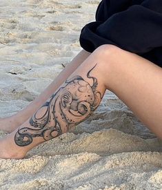 a woman sitting in the sand with an octopus tattoo on her leg