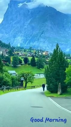 there is a man walking down the street in front of mountains and houses with trees on both sides