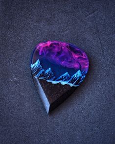 a purple and blue guitar pick sitting on top of a black surface with mountains in the background