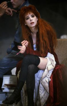 a woman with long red hair sitting on a couch next to a man in black