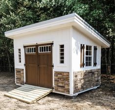 a small white shed with brown doors and windows on the side, sitting in front of some trees