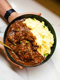 two hands holding a bowl filled with meat and mashed potatoes