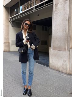 a woman standing in front of a building wearing jeans and a blazer with her hands in her pockets