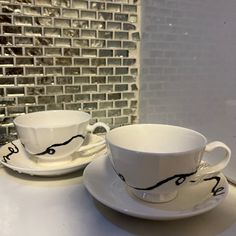 two white cups and saucers with black designs on them sitting on a counter top