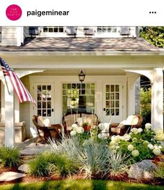 an american flag is on the porch next to some chairs and flowers in front of a house