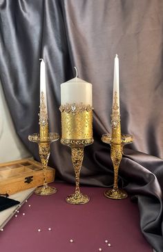 two candles are sitting next to each other on a purple tableclothed surface with silver and gold decorations