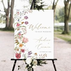 a welcome sign is decorated with flowers and greenery on an easel in front of trees