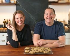 a man and woman sitting at a table with cookies in front of them on a cookie sheet