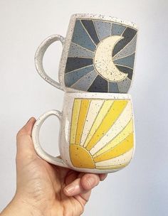 a hand holding two coffee mugs with the sun and moon painted on each one