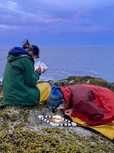 two people are sitting on the rocks by the ocean reading books and eating food from plates