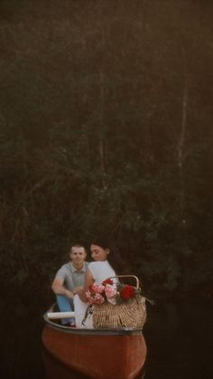 a man and woman sitting in a boat with flowers on the front, surrounded by trees