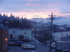 cars parked on the side of a road near buildings and mountains in the background at dusk