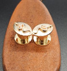 two gold cufflinks on top of a wooden surfboard with a snake in the middle