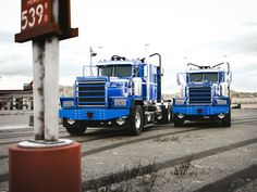 two blue semi trucks parked next to each other on the road in front of a sign