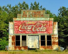 an old coca - cola building in the middle of a field with trees behind it