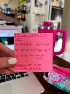 a hand holding a pink note with writing on it in front of a laptop computer