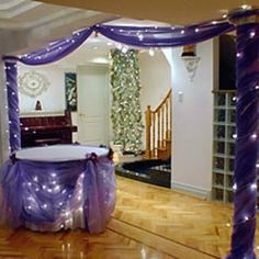 a room decorated in purple and white with lights on the ceiling, table cloth draped over it