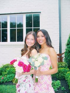 two young women standing next to each other in front of a white building holding flowers