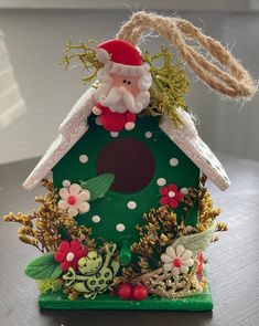 a green birdhouse with santa clause on top and flowers around the base, sitting on a table