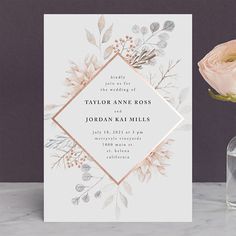 a white and gold wedding card next to a glass vase with a flower in it