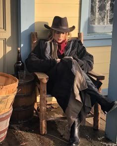 a woman sitting on a wooden chair next to a barrel and a bottle in front of a house