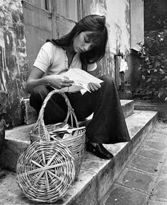 a woman sitting on the steps reading a book and holding a wicker basket next to her