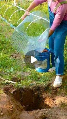 a woman in overalls and pink shirt holding a blue bucket filled with dirt next to a hole