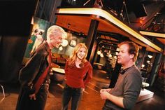 two men and a woman standing in front of a television screen talking to each other