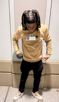 a young person with dreadlocks standing in front of a wall wearing a tan shirt and black pants