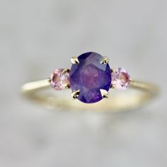 Why I Love It: Crowd Pleaser is sure to be a Gem Breakfast fan favorite! At the center sits a 1.16 carat medium oval cut opalescent sapphire that looks positively effervescent. On either side sit a pair of vibrant pink round brilliant cut sapphires that just add to the uniqueness of this ring. topped with a serving of 14k Yellow Gold and held by stunning claw prongs, you don't get closer to the perfection than this. The Details: 14k Yellow Gold 1.16 Carat Purple Oval Cut Opalescent Sapphire Sri Luxury Purple Oval Sapphire Ring, Ring Inspo, Claw Prong, Purple Sapphire