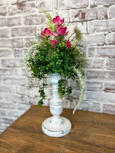 a white vase with pink flowers and greenery on a wooden table next to a brick wall