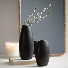 two black vases with white flowers in front of a mirror and candle on a table