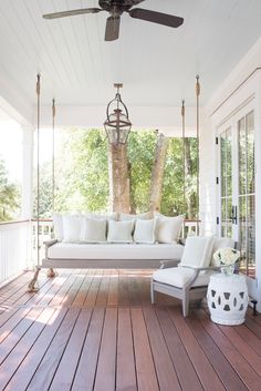 a porch swing with white cushions and pillows on the front porch, hanging from a ceiling fan