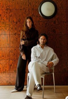 a man and woman are sitting on chairs in front of a wall with wood paneling