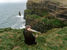 a person sitting on top of a grass covered hill next to the ocean with cliffs in the background