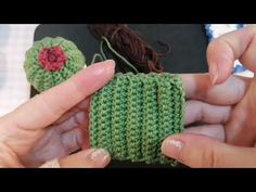 two hands are holding small crocheted items in green and red colors, one has a flower on it