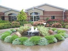 a fountain surrounded by plants and flowers in front of a brick building with large windows