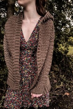 a woman standing in front of some trees wearing a brown cardigan and floral dress