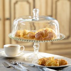 two croissants on a glass cake stand with a cup of coffee and silverware