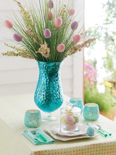 a blue vase filled with pink flowers on top of a green table cloth next to cups and utensils