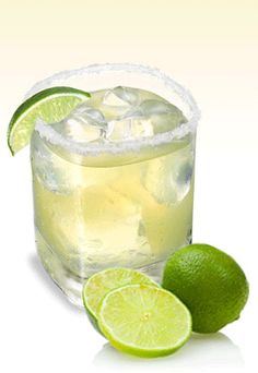 two limes next to a glass of lemonade with ice and lime slices on the rim