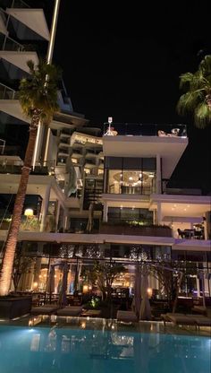 an outdoor swimming pool with palm trees in front of a building at night time,