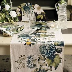 a table topped with a white and blue flower covered table cloth next to a vase filled with flowers