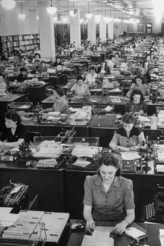 women working at desks in an office with lots of papers and telephones on them