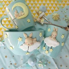 two mugs decorated with bunnies and stars are on a blue tablecloth next to other items