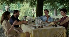 four people sitting at a table in the shade under a tree, eating and drinking