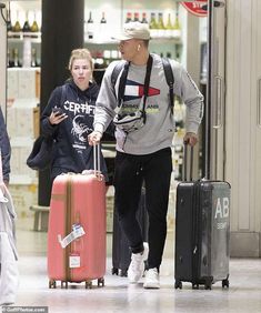 a man and woman walking through an airport with luggage on their back, one carrying a pink piece of luggage while the other is using her cell phone