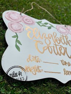 a white sign with gold lettering on it sitting in the grass next to some flowers